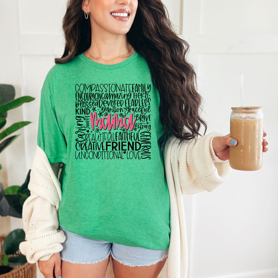 Mama Word Collage DTF Heat Transfer, colorful design on a green T-shirt with positive words such as 'kind', 'brave', 'love', and 'daughter' in various fonts, worn by a smiling woman holding a cup of coffee.