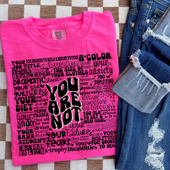You Are Not Polka Dots DTF Heat Transfer - vibrant pink t-shirt with black typographic design depicting empowering messages, laid flat beside ripped jeans on a checkered background.
