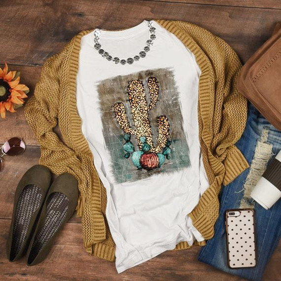 Leopard Cactus Sublimation Transfer, a graphic T-shirt featuring a painted cactus with leopard print, laid out on a rustic wooden surface with casual fashion accessories around it.