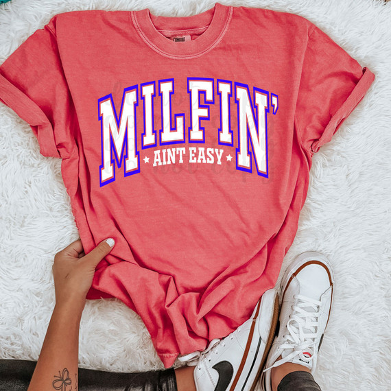 Milfin' Aint Easy DTF Heat Transfer, pink graphic t-shirt with bold 'MILFIN' Ain't Easy' text, on a white fluffy background with a person wearing white sneakers.