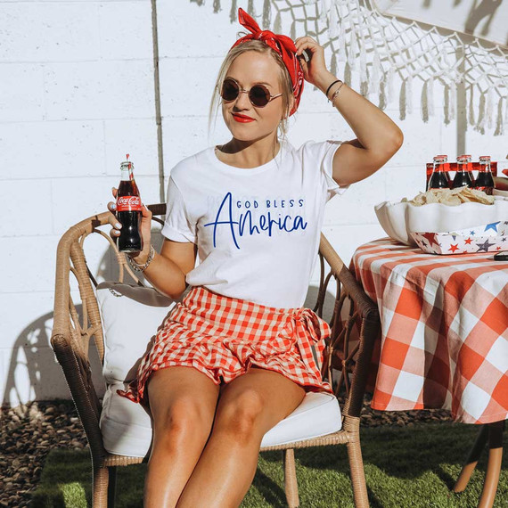God Bless America Blue DTF Heat Transfer, patriotic woman at outdoor celebration wearing white T-shirt with 'God Bless America' print and red gingham shorts, holding a Coca-Cola bottle.
