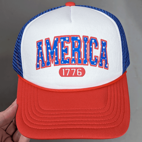America 1776 PUFF Hat/Pocket Screen Print Heat Transfer, close-up view of a patriotic trucker cap with a red brim, blue mesh back, and a white front panel featuring bold 'America' text with stars and '1776' underneath.
