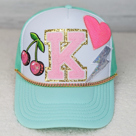 Pink Enamel Gold Hat Chain, colorful hat with patches including a pink letter K, cherries, a pink heart, and a lightning bolt, featuring a gold chain embellishment.