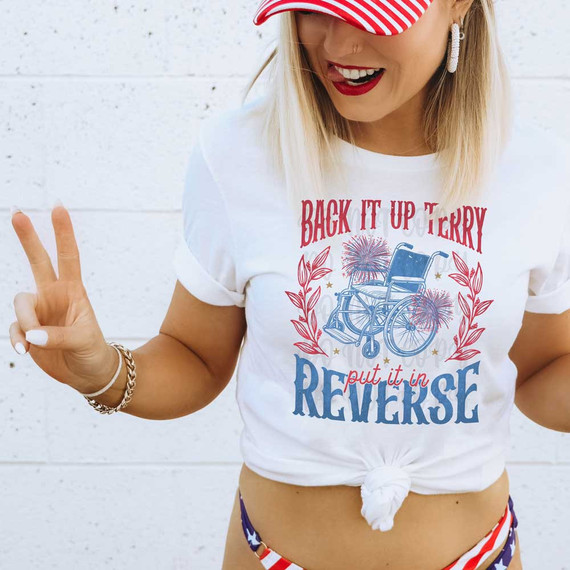 Back It Up Terry DTF Heat Transfer, image of a woman wearing a white T-shirt with a graphic of a wheelchair surrounded by fireworks and text, knotted at the waist, with red lipstick and patriotic accessories.