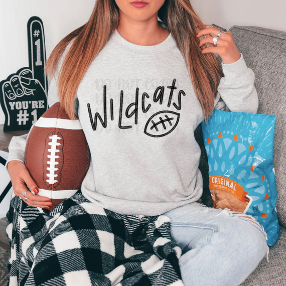 Wildcats Football DTF Heat Transfer, woman wearing a sweatshirt with Wildcats football design, holding a football, sitting on a couch with snacks and decorations in the background.