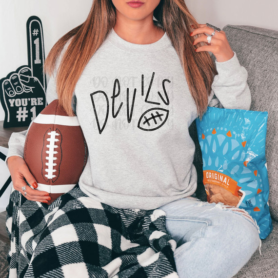 Devils Football DTF Heat Transfer, woman wearing a gray sweatshirt with a hand-drawn devil design and text, sitting on a couch with football and snack bag.
