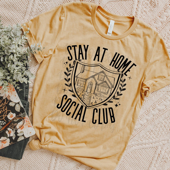 Stay At Home Social Club DTF Heat Transfer, image of a beige T-shirt with a black graphic and text design for a social club, laid on a textured pink blanket with a plant and floral gift bag nearby.