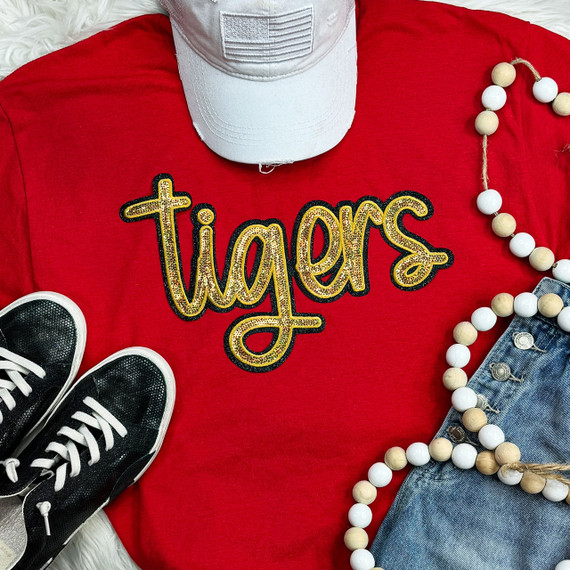Tigers Gold Sequin Patch, close-up view of a decorative golden sequin embroidered patch featuring the word 'tigers' on a red fabric shirt, with accessories including black sneakers, white cap, wooden bead necklace, and denim jeans.