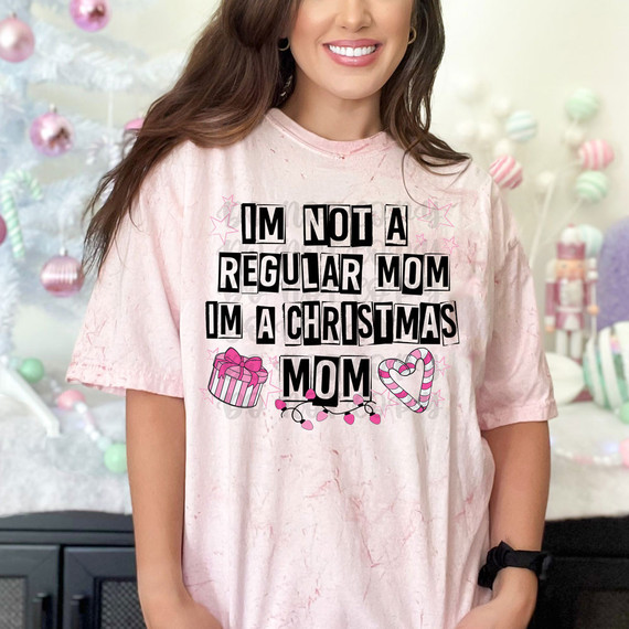 Product Name, I Am Not A Regular Mom I'm A Christmas Mom DTF Heat Transfer on a pink t-shirt worn by a smiling woman in holiday decorated background.