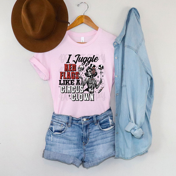 I Juggle Red Flags Like A Circus Clown DTF Heat Transfer, pink t-shirt with circus-themed skeleton and clown design, surrounded by red flags, casual denim shorts, a denim shirt, and a brown hat hanging on a wall.