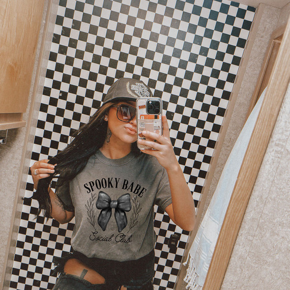 Spooky Babe Social Club DTF Heat Transfer, woman taking a mirror selfie wearing a gray T-shirt with the design of a black bow and the words 'Spooky Babe Social Club' on it.