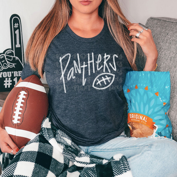 Panthers Football WHITE DTF Heat Transfer, close-up of a woman wearing a dark gray t-shirt with 'Panthers' written in white with a football graphic, holding a football, on a gray sofa, with blue snack bag nearby.