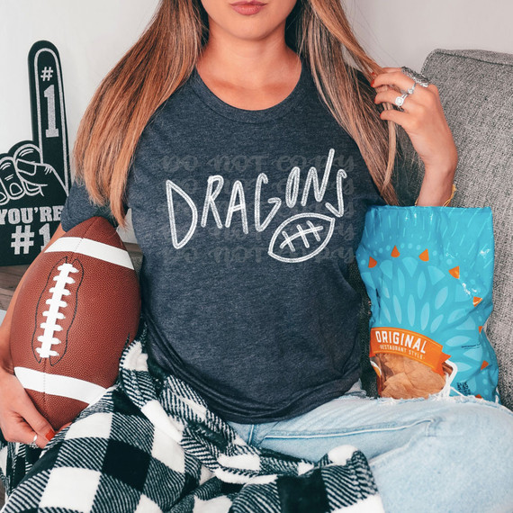 Dragons Football WHITE DTF Heat Transfer, a white heat transfer design featuring the word 'Dragons' and a football graphic, displayed on a gray T-shirt worn by a woman holding a football and a bag of snacks.