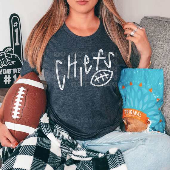 Chiefs Football WHITE DTF Heat Transfer, image of a woman wearing a Chiefs football-themed T-shirt with a football graphic, holding a football, and sitting beside a bag of chips.