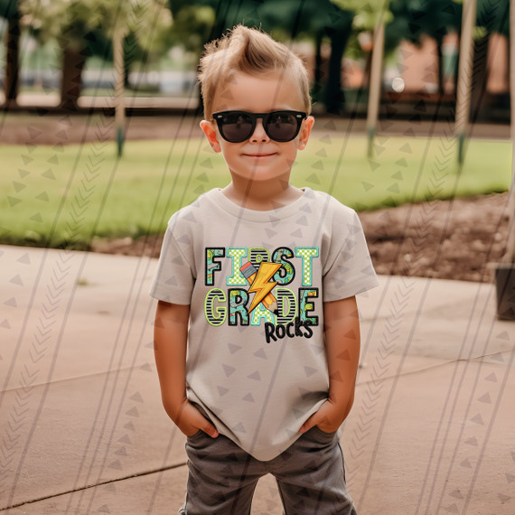 First Grade Rocks Blue/Green DTF Heat Transfer, colorful design with a lightning bolt, stars, and playful fonts on a child's t-shirt.