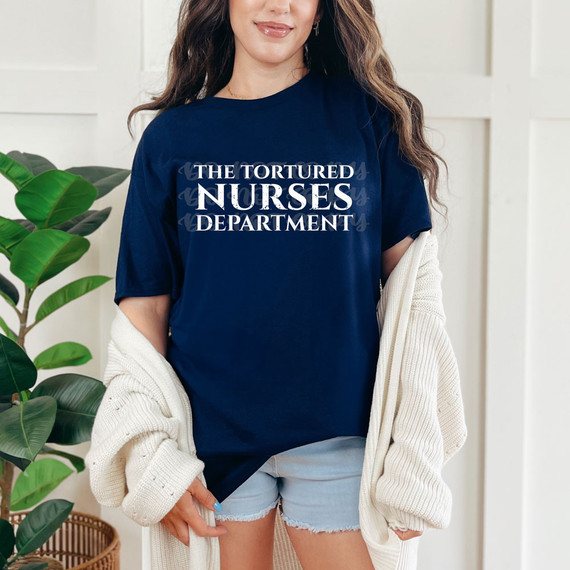 The Tortured Nurses Department DTF Heat Transfer, woman wearing a navy blue T-shirt with white text, casual outfit, indoor setting, green plants in background.
