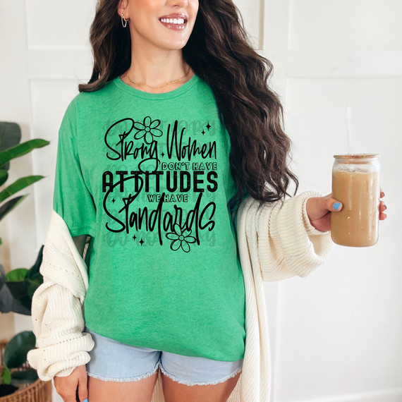 Strong Women Don't Have Attitudes They Have Standards DTF Heat Transfer, a woman wearing a green t-shirt with this bold statement printed in black, holding a jar of beverage, showcasing a fashionable casual style.