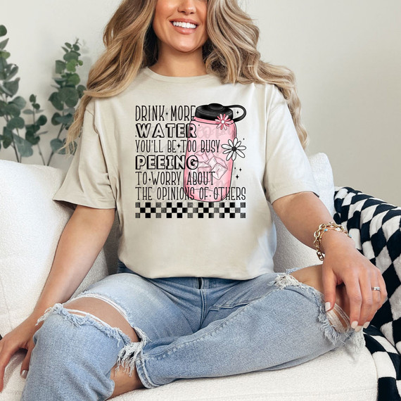 Drink More Water You'll Be Too Busy Peeing To Worry About The Opinions Of Others DTF Heat Transfer T-shirt, a woman sitting on a sofa wearing the humorous graphic T-shirt with a pink water bottle design and playful text.