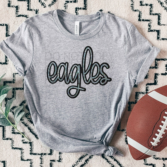 Eagles Faux Sequin Patch DTF Heat Transfer, grey t-shirt with sequin eagle patch design on front, laid on patterned surface near football and green leaves.