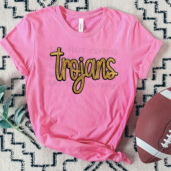 Trojans Faux Gold Sequin Patch DTF Heat Transfer, pink T-shirt with gold sequin patch spelling 'Trojans' on the front, placed on a textured background with a football nearby.