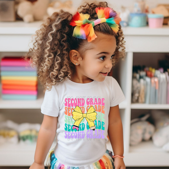 Second Grade Coquette Pencil Bow DTF Heat Transfer, colorful and playful children's design on a white t-shirt worn by a young girl with curly hair and a rainbow bow.