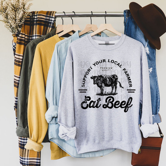 Support Your Local Farmer Eat Beef DTF Heat Transfer, graphic T-shirt with a cow illustration displayed among other casual clothing and accessories.