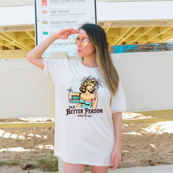 I'm A Better Person When I'm Tan DTF Heat Transfer, a woman wearing a white t-shirt with a retro woman illustration and the text 'I'm A Better Person When I'm Tan' at the beach.