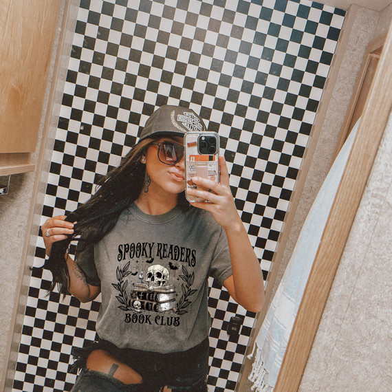 Spooky Readers Book Club DTF Heat Transfer, a woman wearing a gray T-shirt with a spooky book-themed graphic including a skull and stacked books, taking a mirror selfie in a checkered wall bathroom.