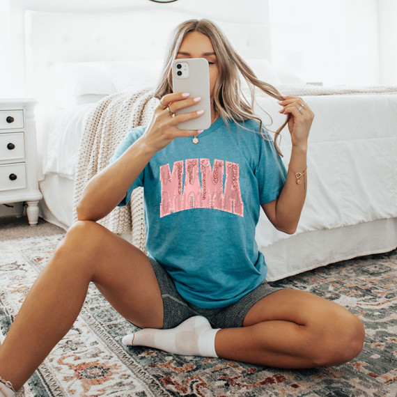 Mama Faux Glitter Drip DTF Heat Transfer, a close-up of a woman wearing a blue t-shirt with pink glitter drip design, sitting on the floor in a bright bedroom while taking a mirror selfie.