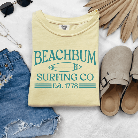 Beach Bum Surfing Co DTF Heat Transfer, yellow T-shirt with surfboard graphic and text, surrounded by sunglasses, denim shorts, slippers, and tropical leaves.
