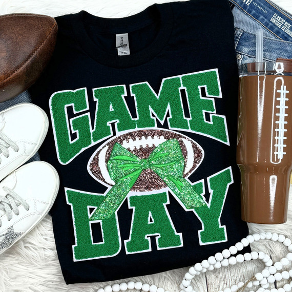 Green Chenille Football Game Day with Sequin Bow Patch, close-up of a black T-shirt featuring a glittery green bow on an American football, surrounded by white sneakers, a brown tumbler, and layered white bead necklaces, perfect for sports or football themed occasions.