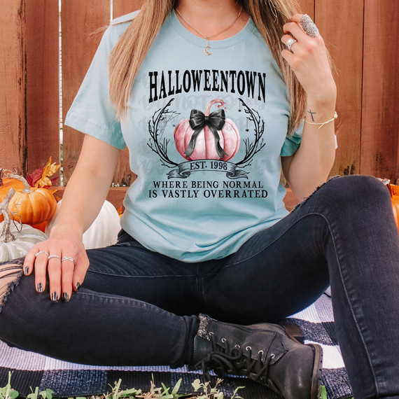 HT Coquette Pumpkin DTF Heat Transfer, pink pumpkin with black bow design with Halloween-themed text on a light blue T-shirt worn by a woman outdoors.