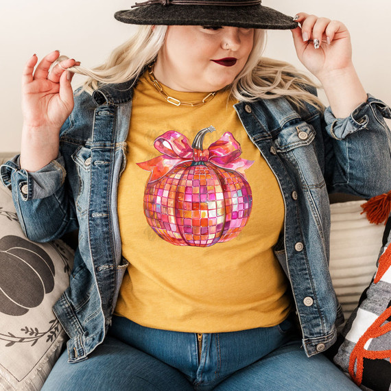 Disco Pumpkin DTF Heat Transfer, a colorful, reflective pumpkin graphic with a bow and metallic design on a yellow T-shirt worn by a woman in a denim jacket and black hat.