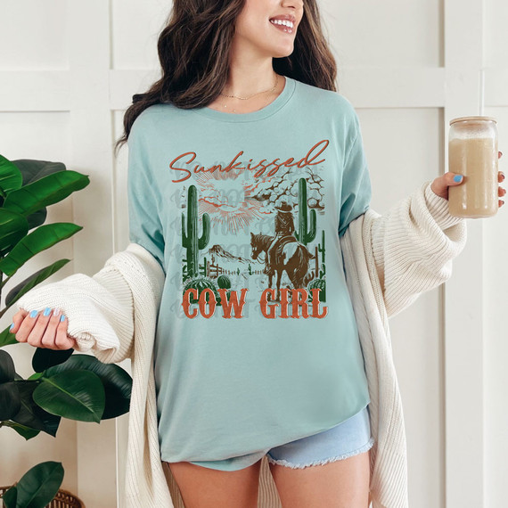 Sunkissed Cow Girl DTF Heat Transfer, a woman wearing a light blue t-shirt with cowgirl and desert scene graphic, holding a drink, emphasizing the product's design and vibrant colors.