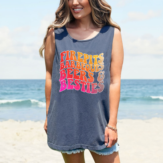 Firepits Barbeques Beers & Besties DTF Heat Transfer T-shirt, a woman wearing a sleeveless blue T-shirt with colorful text, standing on the beach with the ocean in the background.