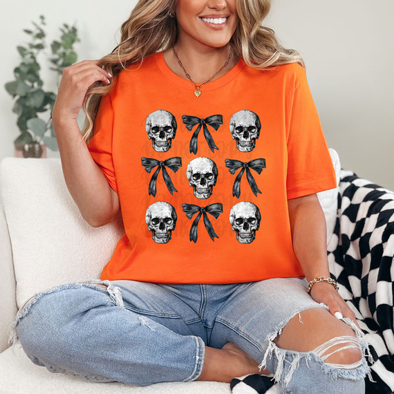 Skull Coquette DTF Heat Transfer, a vibrant orange T-shirt with skull and bow design, worn by a smiling woman sitting on a cozy couch.