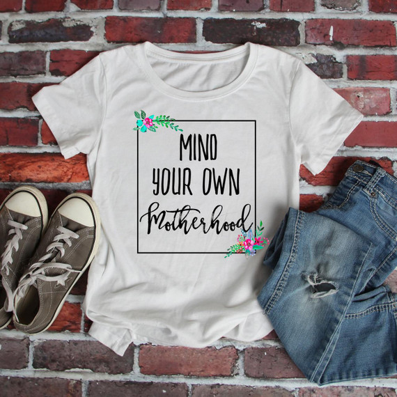 Mind your own motherhood sublimation transfer, white T-shirt with floral accents and motivational quote, laid out with casual sneakers and ripped jeans on a brick wall background.