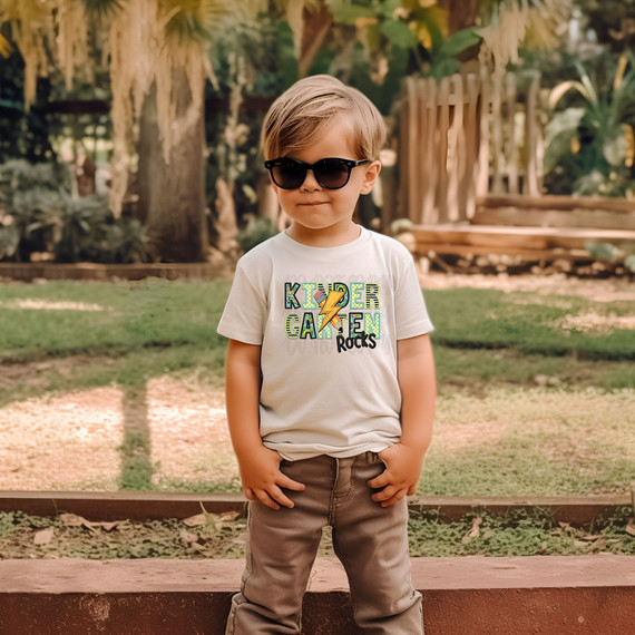 Kindergarten Rocks Blue/Green DTF Heat Transfer, a young boy wearing sunglasses and a t-shirt with the design, standing outdoors in a park.