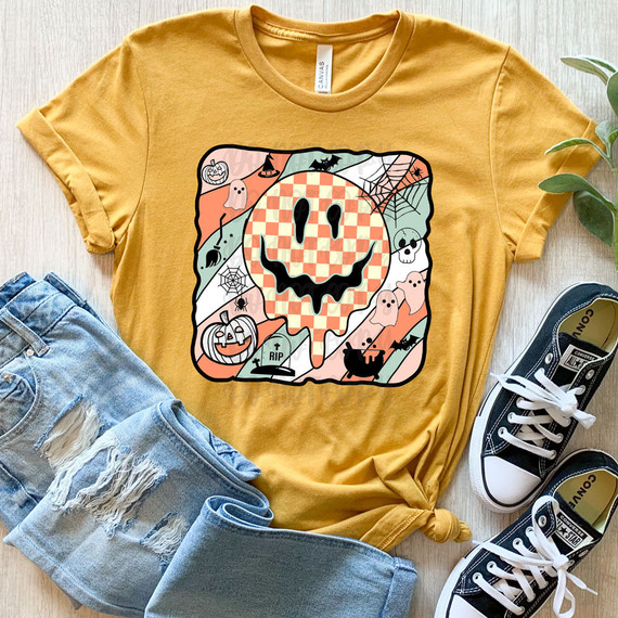 Spooky Happy Face DTF Heat Transfer, Halloween-themed design on a yellow t-shirt featuring a smiling pumpkin face surrounded by ghosts, bats, spider webs, and tombstones.