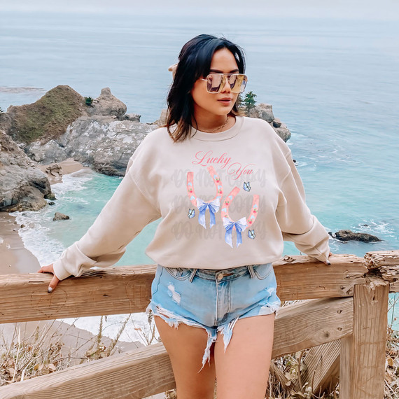 Lucky You DTF Heat Transfer, a colorful design of two handbags with floral patterns and bows, displayed on a woman's sweatshirt in a scenic beach setting.