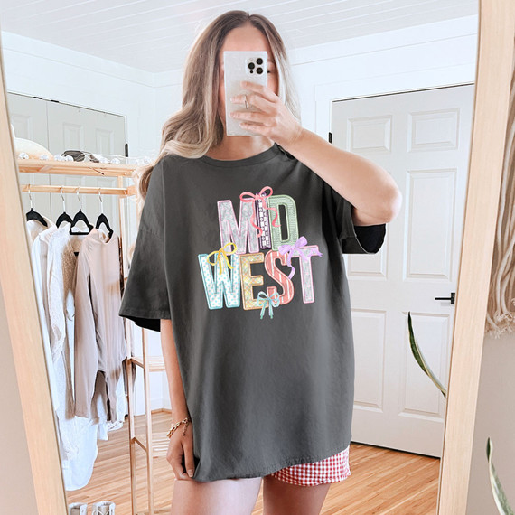 Midwest Coquette Faux Embroidery DTF Heat Transfer, image of a person wearing a black oversized t-shirt with colorful decorative letters spelling 'MIND WEST' with bows, taking a mirror selfie in a bedroom.