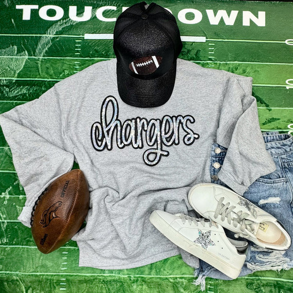 Chargers Silver Sequin Patch, a casual outfit including a gray sweatshirt with 'chargers' embroidered in silver sequins, a black baseball cap with football patch, white sneakers with star design, distressed denim shorts, and a brown football, laid out on a green painted wooden surface.