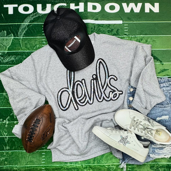 Devils Silver Sequin Patch, a shiny sequin patch featuring the word 'devils' in a stylized script on a light gray sweatshirt surrounded by sneakers, a brown football, a black cap with football embroidery, and casual denim shorts on a green textured background.