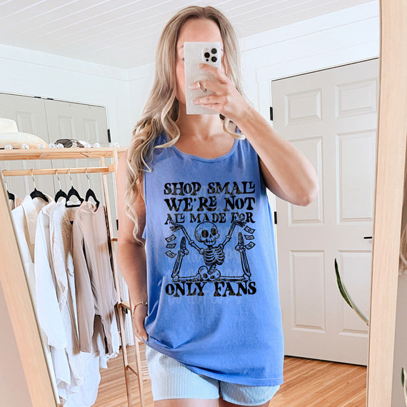 Shop Small We're Not All Made For Only Fans DTF Heat Transfer, woman wearing a blue sleeveless shirt with black text and a skeleton graphic, taking a mirror selfie in a room with wooden floors and clothing on a rack.