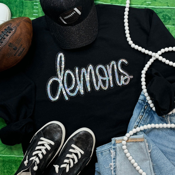 Demons Sequin Patch, an embroidered patch with shinny, sparkling sequins spelling 'demons' on a black sweatshirt, surrounded by casual fashion accessories including sneakers, jeans, a pearl necklace, a cap, and an American football on a green background.