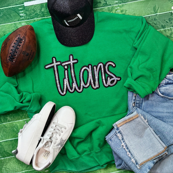 Titans Sequin Patch, a clothing item featuring a green sweatshirt with a sequined 'titans' patch, paired with white sneakers, denim jeans, a black cap, and a vintage football on a green wooden background.