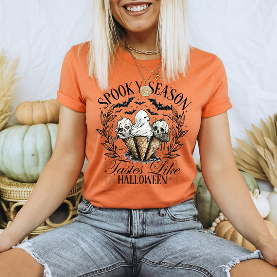 Spooky Season Tastes Like Halloween DTF Heat Transfer, a woman wearing an orange graphic T-shirt with for Halloween themed design featuring skulls and ice cream cones.