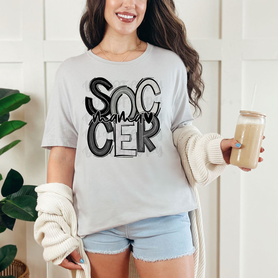Soccer Mama Monochrome DTF Heat Transfer, a woman wearing a white t-shirt with a black and white soccer-themed design, holding a jar drink, in a cozy indoor setting.