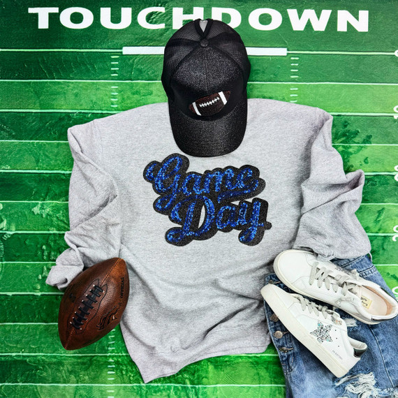 Royal Blue Sequins Game Day Patch, a sparkling blue sequins patch with the words 'Game Day' embroidered on a gray sweatshirt, paired with white sneakers, a black football cap, and a football on a green football field background.