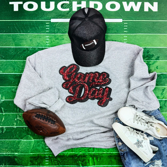 Red Sequins Game Day Patch, a decorative patch with shiny red sequins spelling 'Game Day' on a sweatshirts, displayed alongside a football, a gray cap with a football logo, white sneakers, and jeans, laid out on a green football field background.